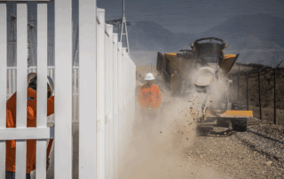 Construction workers operate heavy equipment while installing a rock barrier alongside a white utility fence, with dust and debris flying as material is discharged.